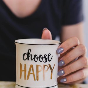 A woman holds a mug with 'choose happy' text indoors, promoting positivity.