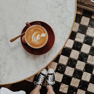Overhead view of a latte with art on a marble table, sneakers visible on checkered floor.