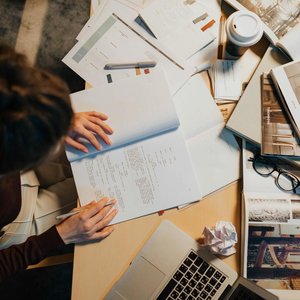 Overhead view of a cluttered work desk with a person writing notes, surrounded by papers, laptop, and coffee cup.