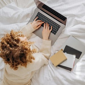 Woman with curly hair typing on a laptop in a cozy bedroom, surrounded by notebooks.