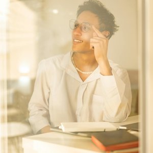 Man with glasses sitting indoors, pensive and relaxed. Clearly reflecting through a window with warm lighting.
