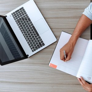 Person writing notes at desk with a laptop and open notebook, capturing a modern workspace.