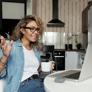 Smiling woman with glasses having a video call from home, enjoying a cup of coffee in her kitchen.