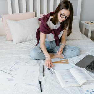 A young woman studying on her bed with a laptop, books, and papers scattered around.