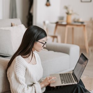 A young woman sits on the floor using a laptop, working remotely in a cozy living room.
