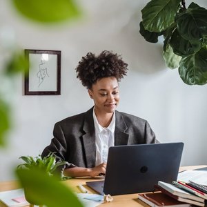 A young woman in a suit working on a laptop at a home office surrounded by plants.