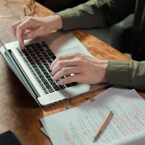 Focused person typing on laptop with notes and papers beside on wooden table.
