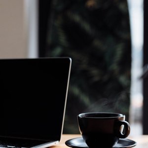 A steaming cup of coffee sits next to a laptop on a wooden desk indoors.