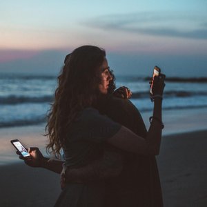 Couple embracing on a beach at sunset, using mobile phones, symbolizing modern love.