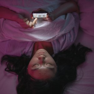 A woman lying in bed at night browsing her smartphone under soft lighting.