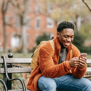 A cheerful young man in warm clothes smiling while using his phone on a bench in a city park.