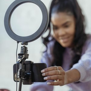 Cheerful young African American female blogger in stylish sweater smiling while setting up camera of smartphone attached to tripod with ring light before recording vlog