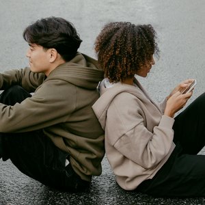 A diverse young couple sits back to back using their smartphones, depicting modern communication.