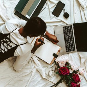 African woman working remotely on laptop with notebooks and flowers around. Comfortable home setup.