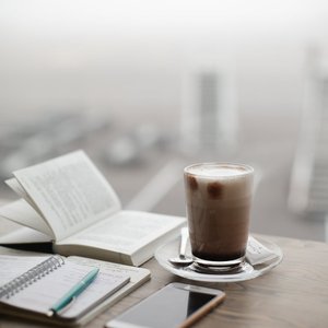 A warm coffee sits beside a notebook and smartphone on a foggy day.
