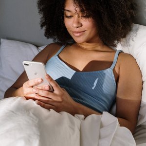 African American woman enjoying a cozy morning in bed, using a smartphone.
