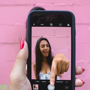 A woman captures her selfie using a smartphone against a vibrant pink wall.