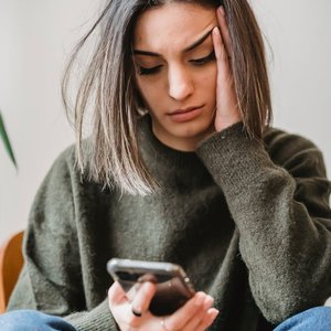 Unhappy female in casual wear leaning on hand and surfing internet on cellphone while sitting in light room near wall at home