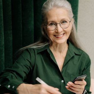 Smiling senior woman with gray hair and glasses holding a smartphone indoors.