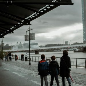 A family walks along the riverbank in Bordeaux on a cloudy day, with cruise ships in the distance.