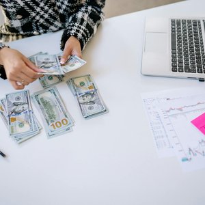 Person counting cash next to laptop and stock market charts on a white table.