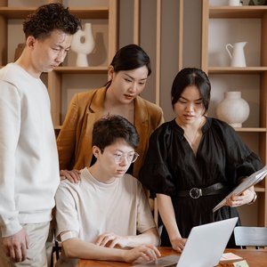 A group of Asian professionals engaged in collaborative work around a laptop in an office setting.