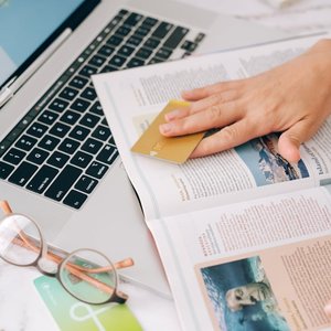 Person holding a credit card while reading a magazine next to a laptop.
