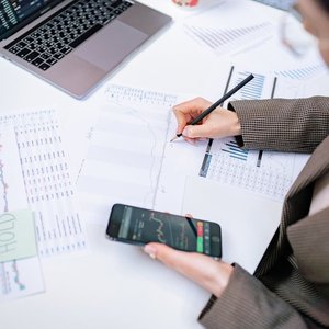 Adult woman analyzing stock market data with laptop and smartphone in office setting.