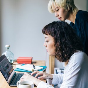 Side view of young women wearing casual clothes working on project using laptop while working on project together and drinking coffee