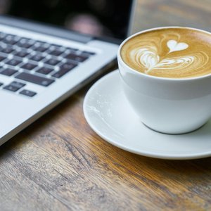 A cup of latte with intricate art beside a laptop on a wooden table, depicting a cozy work setting.