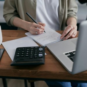 A woman writes financial calculations in a notebook, using a calculator and laptop at a wooden desk.