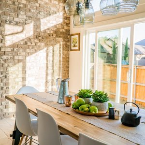 Sunlit dining room with contemporary decor and a rustic brick wall, featuring stylish lighting and a wooden table.