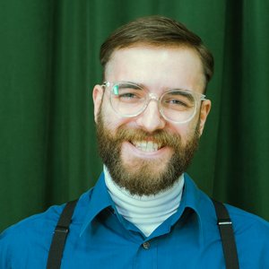 Portrait of a smiling man with beard and eyeglasses against a green curtain background.