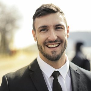 A confident businessman smiling in a black suit with an outdoor background.