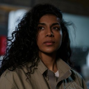 A thoughtful portrait of a woman with curly hair in an industrial setting, captured indoors with dramatic lighting.