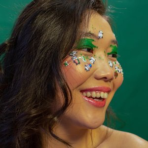 Asian woman smiling with green eye makeup and playful face stickers against a green background.