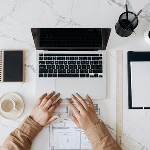 Top view of a stylish home office desk with a laptop, planner, and coffee cup, showing hands on a blueprint.