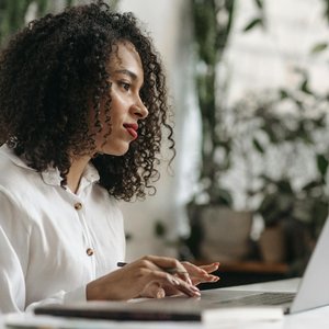 Young woman with curly hair typing on laptop in a bright, plant-filled office space.