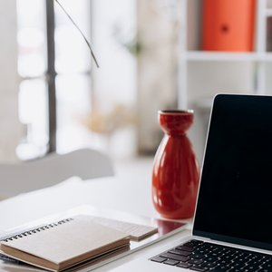 A minimalist home office setup featuring a laptop, notebook, and a red vase on a white desk.