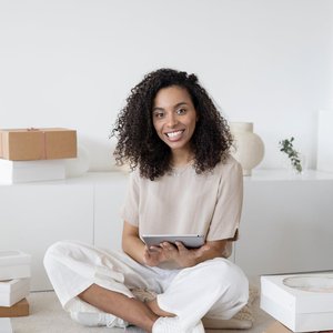 A confident young woman sitting among packages and papers while using a tablet, showcasing modern entrepreneurship.