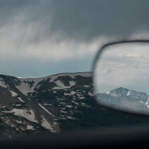 Snow-capped mountains reflected in a rearview mirror with dramatic cloudy skies.