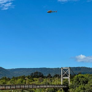 A helicopter flies above a suspension bridge in Fox Glacier, New Zealand, under a bright blue sky.