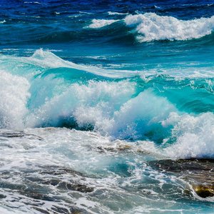 Vibrant blue ocean waves crashing against rocky shoreline under clear summer skies.