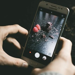 Close-up of a smartphone photographing chocolate pieces and raspberries on a dark surface.
