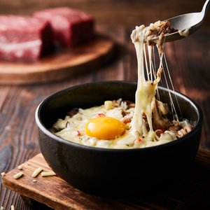A delicious melted cheese and egg bowl on a wooden board with beef in the background.