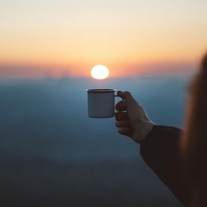 A person holds a coffee cup against a beautiful sunrise, capturing a serene outdoor moment.